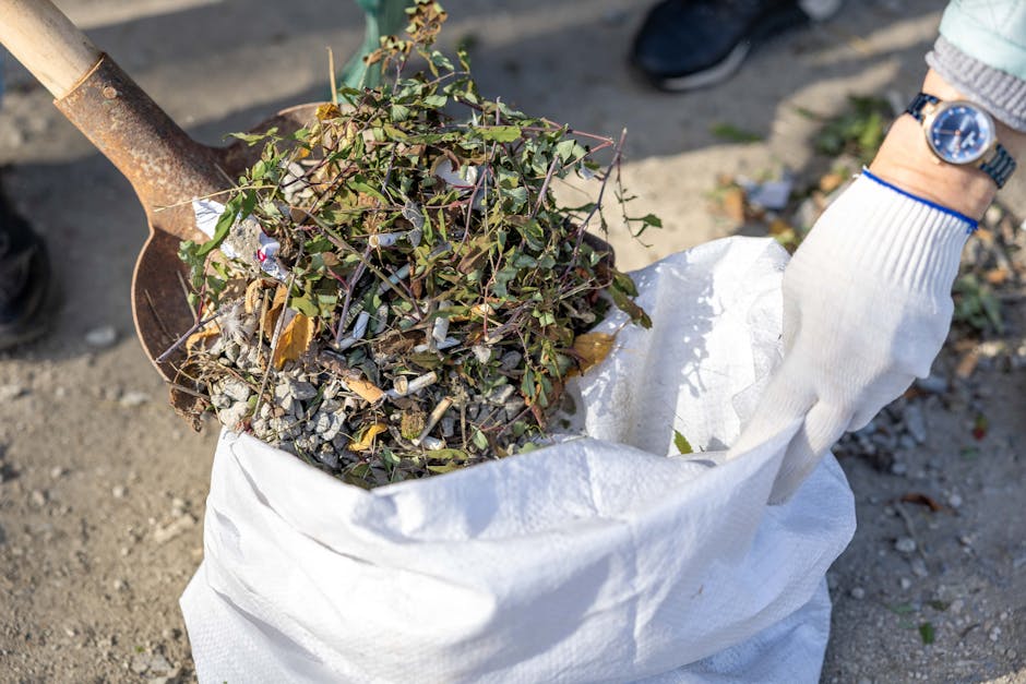 A person wearing a white glove is holding an open white rubbish bag filled with garden debris, including small branches, leaves, and gravel. The debris features a mix of green and brown foliage, with some leaves and twigs scattered among grey gravel. The bag is positioned on a gravelled surface, and a spade with a rusty metal blade is partially visible, leaning into the bag. The background shows a paved area with a person's black shoe and some indistinct objects, suggesting an outdoor setting suitable for garden waste collection or site clearance activities. The scene reflects a typical outdoor rubbish removal effort, possibly conducted by a private waste disposal service such as Rubbish Clearance Brent, to manage garden waste and non-toxic debris in preparation for disposal or recycling.