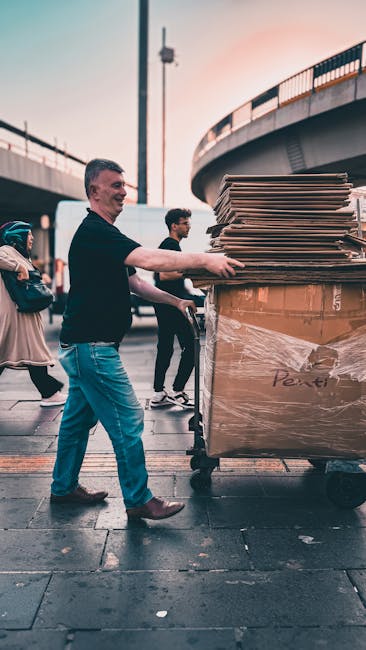 A man with short dark hair, dressed in a black shirt and light blue jeans, is standing outdoors during early evening under an elevated road or bridge structure. He is pushing a large, black trolley loaded with a substantial pile of flattened cardboard boxes, stacked neatly and partially wrapped in clear plastic. In the background, two other individuals, including a person with curly hair and glasses, are walking nearby. The sky shows a soft gradient of light, indicating dusk, and the environment appears to be an urban setting with paved surfaces and metal railings supporting the bridge above. The scene captures a moment of independent waste handling or rubbish clearance, with the cardboard likely collected for disposal or recycling, aligning with private waste management services that offer alternative rubbish removal solutions. Rubbish Clearance Brent specializes in rubbish, and this scene exemplifies on-site clearance of bulky waste materials, emphasizing manual loading and the use of equipment suited for handling large cardboard loads in an outdoor, city landscape.