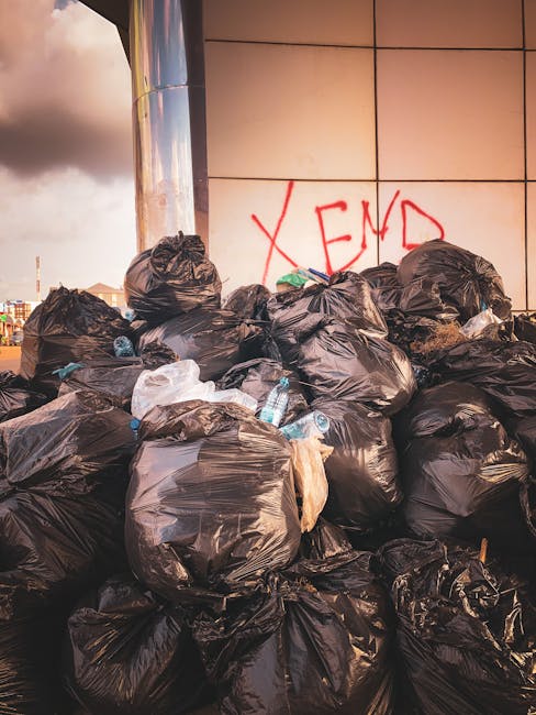 A large pile of black plastic rubbish bags filled with waste is stacked against a tiled wall, with some partially torn or open, revealing plastic bottles and assorted debris inside. The bags vary in size and are situated on a paved surface, possibly a street or alleyway, under diffuse natural lighting that suggests late afternoon or early evening. In the background, part of a clean, modern building with a reflective metal surface and beige tiles is visible, along with a graffiti tag in red spray paint reading 'XEND' on the wall. The overall scene depicts an accumulation of waste awaiting collection, indicating the need for rubbish removal services such as those offered by Rubbish Clearance Brent, which specializes in private waste clearance for commercial and residential clients.