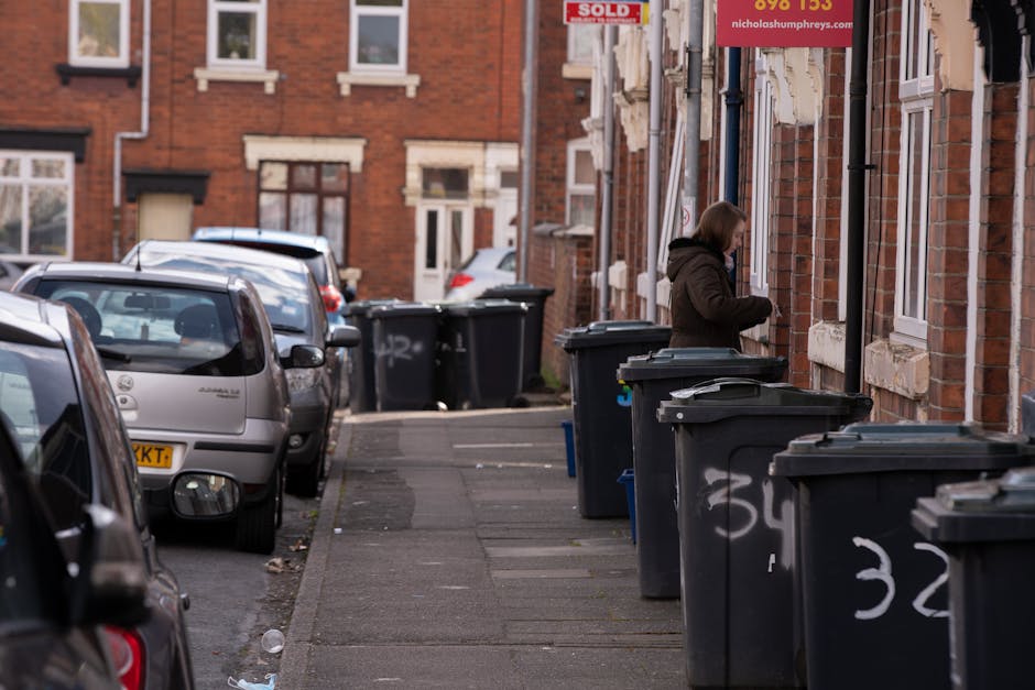 A residential street with a row of black wheelie bins lined up along the pavement in front of brick terraced houses. The bins are standard size, made of plastic, with hinged lids, and some are marked with white spray paint numbers. A woman dressed in a dark brown hooded jacket is standing beside one of the bins, appearing to be in the process of opening or closing it. The street contains several parked cars, including a silver hatchback at the forefront and others further down, with visible rear and side windows, bumpers, and mirrors. The background shows additional brick houses with white-framed windows, some with decorative architectural details such as window sills and lintels. The natural lighting suggests daytime, with soft shadows indicating an overcast sky. The scene connects to private waste removal services, highlighting an alternative to council rubbish collection, with Rubbish Clearance Brent potentially providing on-site clearance or waste disposal solutions for residents and property owners.