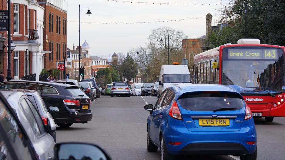 A busy urban street scene in Harlesden NW10 showing parked cars along both sides of the road, including a black and a silver vehicle in the foreground. A blue hatchback with a yellow registration plate is prominent in the center, partially blocking the view of the street. On the right, a red Metroline bus displaying the route number 143 towards Brent Cross is moving past. Behind the vehicles, there are brick terraced houses with white window frames and architectural details, and some trees with sparse foliage indicating early spring or late autumn. The street is illuminated by traditional-style lamp posts with hanging string lights, adding ambient lighting to the scene. Further down the road, additional parked cars and a white van are visible, with a distant view of a church spire and more buildings in the background. The overall atmosphere reflects typical city traffic, which Rubbish Clearance Brent might encounter during private waste collection or on-site clearance services in residential areas like Harlesden, where nearby rubbish removal and disposal services are frequently required.