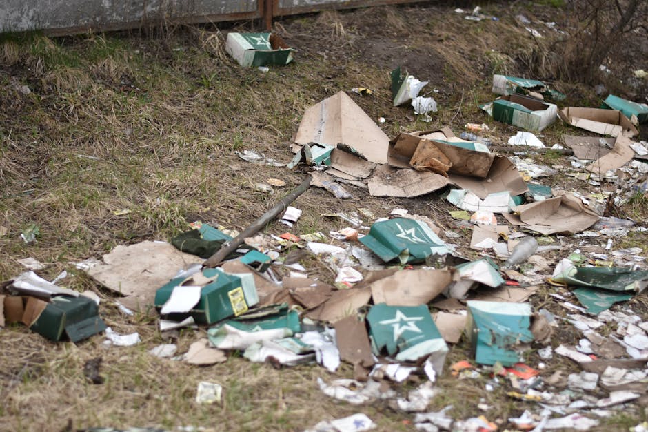 An outdoor scene showing a section of ground with scattered debris consisting of flattened cardboard boxes, some with printed designs in shades of green, white, and beige, along with crumpled paper, plastic bottles, and other waste materials. The boxes are partially crushed, with torn edges and flattened shapes, and are spread across a patch of grass and dirt. In the background, there is a wooden fence with a few shrubs and bushes nearby. The area appears to be an informal dumping site or littered roadside, with no visible signage or waste containers present. The lighting suggests natural daylight, highlighting the uneven textures and the mixture of organic and waste materials on the ground. This scene illustrates the type of rubbish that professional waste removal services, such as Rubbish Clearance Brent, often address for proper disposal or clearance, particularly in an environment that may require independent collection or private waste handling solutions.