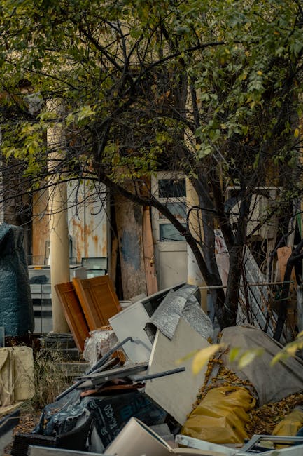 An outdoor scene showing a section of ground with scattered debris consisting of flattened cardboard boxes, some with printed designs in shades of green, white, and beige, along with crumpled paper, plastic bottles, and other waste materials. The boxes are partially crushed, with torn edges and flattened shapes, and are spread across a patch of grass and dirt. In the background, there is a wooden fence with a few shrubs and bushes nearby. The area appears to be an informal dumping site or littered roadside, with no visible signage or waste containers present. The lighting suggests natural daylight, highlighting the uneven textures and the mixture of organic and waste materials on the ground. This scene illustrates the type of rubbish that professional waste removal services, such as Rubbish Clearance Brent, often address for proper disposal or clearance, particularly in an environment that may require independent collection or private waste handling solutions.